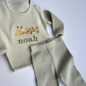 May include: Sage green ribbed knit pajama set. The long-sleeved top features an embroidered giraffe and the name "Noah" in green thread. The set includes matching pants. The fabric has a vertical ribbed texture.