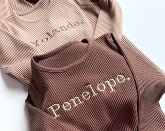 Könnte beinhalten: Zwei gerippte Langarmshirts, eines in Hellbraun und das andere in Dunkelbraun. Der Name "Yolande" ist auf dem helleren Shirt und "Penelope" auf dem dunkleren Shirt gestickt. Die Shirts sind aus weichem Rippmaterial.
