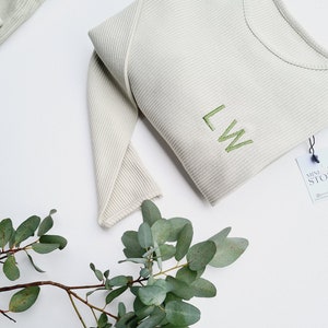 Op de afbeelding: Een lichtgrijs geribd shirt met lange mouwen met de geborduurde initialen "LW" in groen. Een takje eucalyptus met groene bladeren en een label met de tekst "MINI STORE" zijn ook zichtbaar.