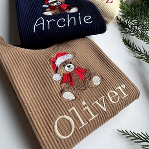 May include: Three ribbed knit shirts in navy blue, tan, and cream. Each shirt features an embroidered teddy bear wearing a Santa hat and scarf, with a name embroidered below. The names are Archie, Oliver, and Willow.