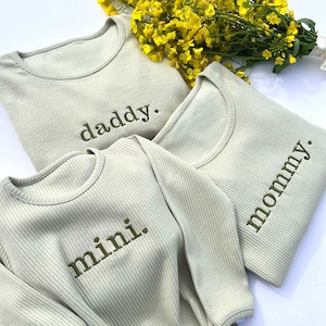 Op de afbeelding: Drie lichtgroene geribde shirts met lange mouwen, geborduurd met de woorden "daddy.", "mommy." en "mini." in olijfgroen. Een boeket gele bloemen staat op de achtergrond.