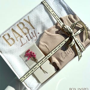 May include: A white blanket with a delicate lace pattern and the words "Baby Lily" embroidered in gold thread. A beige romper with the name "Lily" embroidered on the front is included in the gift box. The box is tied with a ribbon that says "made with love" in black lettering.
