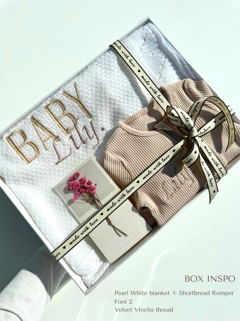May include: A white blanket with a delicate lace pattern and the words "Baby Lily" embroidered in gold thread. A beige romper with the name "Lily" embroidered on the front is included in the gift box. The box is tied with a ribbon that says "made with love" in black lettering.