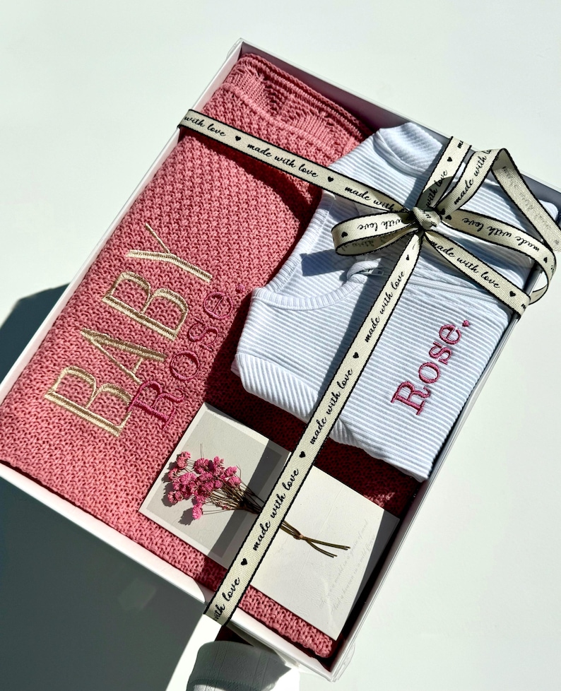 May include: A white gift box with a pink knitted blanket, a white onesie, and a card. The blanket has the words "Baby Rose" embroidered on it. The onesie has the words "Rose" embroidered on it. The ribbon around the box has the words "made with love" printed on it.