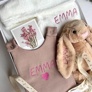 May include: A gift set in a white box, featuring a plush bunny, a bib, and a robe. The bib and robe are embroidered with the name "EMMA" in pink. The bunny is tan with pink accents. A card with pink flowers is also included.