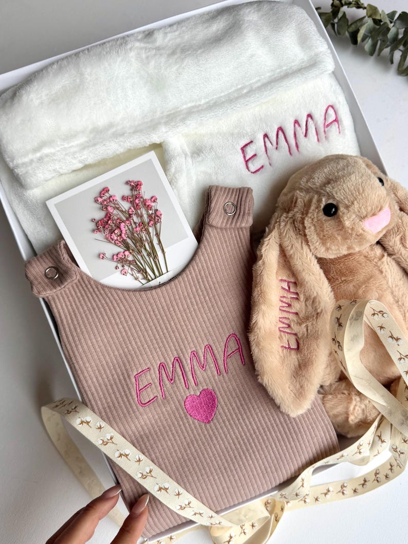 May include: A gift set in a white box, featuring a plush bunny, a bib, and a robe. The bib and robe are embroidered with the name "EMMA" in pink. The bunny is tan with pink accents. A card with pink flowers is also included.
