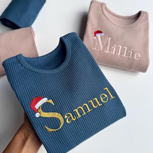May include: Several ribbed knit shirts in shades of blue and pink. One blue shirt has the name "Samuel" embroidered in gold with a Santa hat. Another pink shirt has the name "Millie" embroidered in pink with a Santa hat. The shirts are folded.