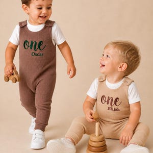 May include: Two toddlers wearing corduroy overalls with the word "one" and a name printed on the front. One is brown, the other is beige. Both wear white t-shirts and trainers. Wooden toys, including a car and stacking rings, are in the image.