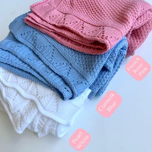 May include: Three knitted baby blankets in different colors: white, light blue, and pink. The blankets are folded and stacked on top of each other. The labels below each blanket identify the colors as Pearl White, CornSilk Blue, and French Rose Pink.
