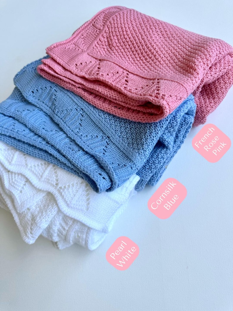 May include: Three knitted baby blankets in different colors: white, light blue, and pink. The blankets are folded and stacked on top of each other. The labels below each blanket identify the colors as Pearl White, CornSilk Blue, and French Rose Pink.