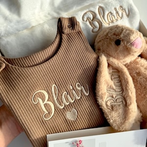 May include: A brown ribbed romper with the embroidered name Blair and a heart, a white plush blanket with the name Blair, a tan plush bunny with the name Blair on its ear, and a white card with pink flowers and the text Especially for you.