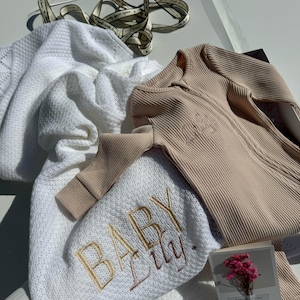 May include: A white knitted baby blanket with the words "Baby Lily" embroidered in gold thread. A beige ribbed baby onesie with the name "Lily" embroidered on the front. A small card with pink flowers.