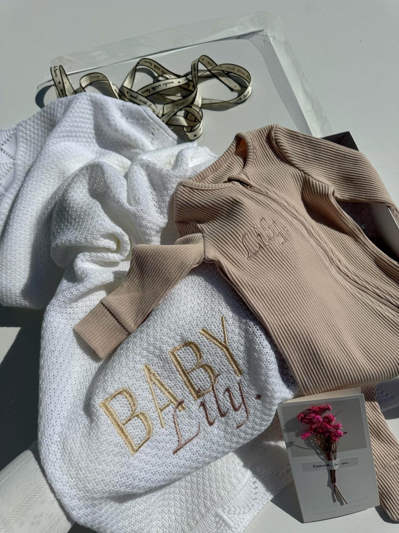 May include: A white knitted baby blanket with the words "Baby Lily" embroidered in gold thread. A beige ribbed baby onesie with the name "Lily" embroidered on the front. A small card with pink flowers.