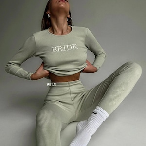 Puede incluir: Una mujer lleva una camiseta de manga larga de color verde claro con el texto "BRIDE" impreso en la parte delantera y unos pantalones de color verde claro a juego. Lleva calcetines blancos con un logotipo de Nike negro en forma de swoosh y tiene el pelo recogido hacia atrás.