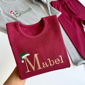 May include: A set of children's ribbed knitwear in gray and burgundy. The burgundy top features the name "Mabel" embroidered in gold with a Santa hat. The gray top has the name "Ethan" embroidered in green with a Santa hat.