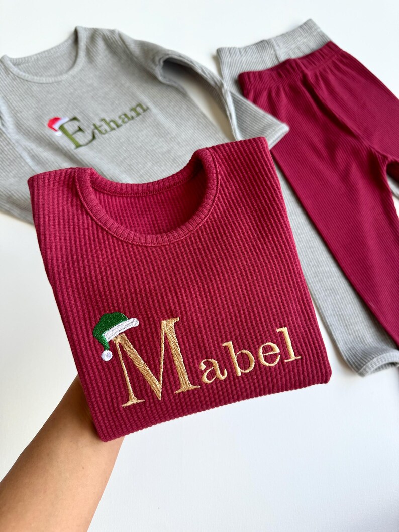 May include: A set of children's ribbed knitwear in gray and burgundy. The burgundy top features the name "Mabel" embroidered in gold with a Santa hat. The gray top has the name "Ethan" embroidered in green with a Santa hat.