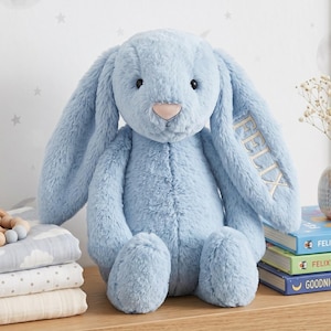 May include: A plush stuffed animal in the shape of a rabbit with soft, light blue fur, black eyes, and a pink nose. The name FELIX is embroidered in light-coloured thread on one of the long, floppy ears. The rabbit is seated in an upright position.