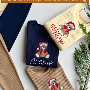 May include: Ribbed knit shirts in navy blue, tan, and cream colors, each embroidered with a festive bear design wearing a Santa hat and scarf. The names "Archie", "Willow", and "Oliver" are embroidered below the bear. The image also shows matching pants.