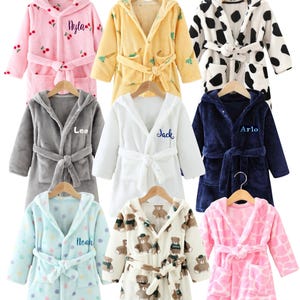 May include: Nine hooded bathrobes in various colors and patterns, including pink with cherries, yellow with leaves, and a black and white cow print. Each robe has a matching belt and the names "Nyla", "Leo", "Jack", "Arlo", and "Noah" embroidered on them.