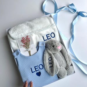 May include: A gift box containing a blue baby outfit, a white plush robe, and a gray stuffed bunny. The outfit and robe are embroidered with the name "LEO" in blue. A small card with flowers is also included.