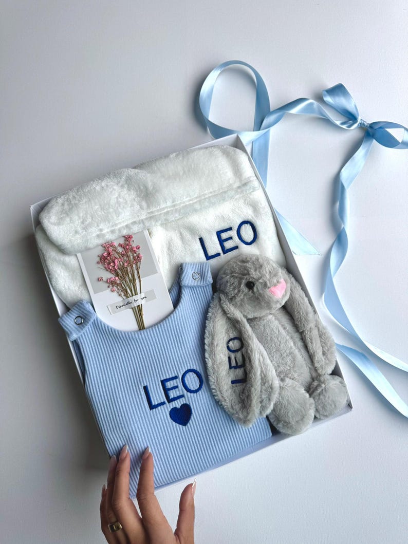 May include: A gift box containing a blue baby outfit, a white plush robe, and a gray stuffed bunny. The outfit and robe are embroidered with the name "LEO" in blue. A small card with flowers is also included.