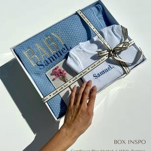 May include: A white gift box with a blue and white baby blanket and a white romper. The box is tied with a blue ribbon with the text "made with love" printed on it. The blanket and romper are embroidered with the name "Samuel".