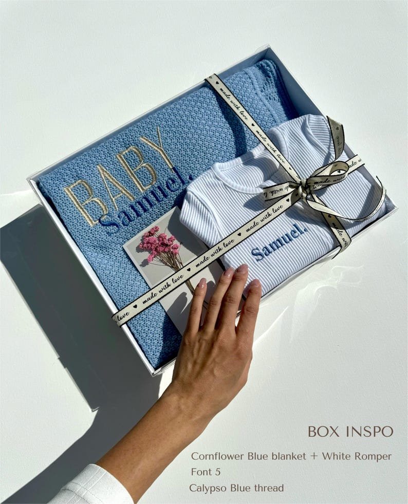 May include: A white gift box with a blue and white baby blanket and a white romper. The box is tied with a blue ribbon with the text "made with love" printed on it. The blanket and romper are embroidered with the name "Samuel".