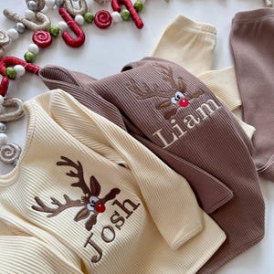 May include: Cream and brown ribbed long-sleeved shirts and pants with embroidered reindeer faces and names. Festive holiday apparel with a Christmas theme. The text "Josh" and "Liam" are visible.