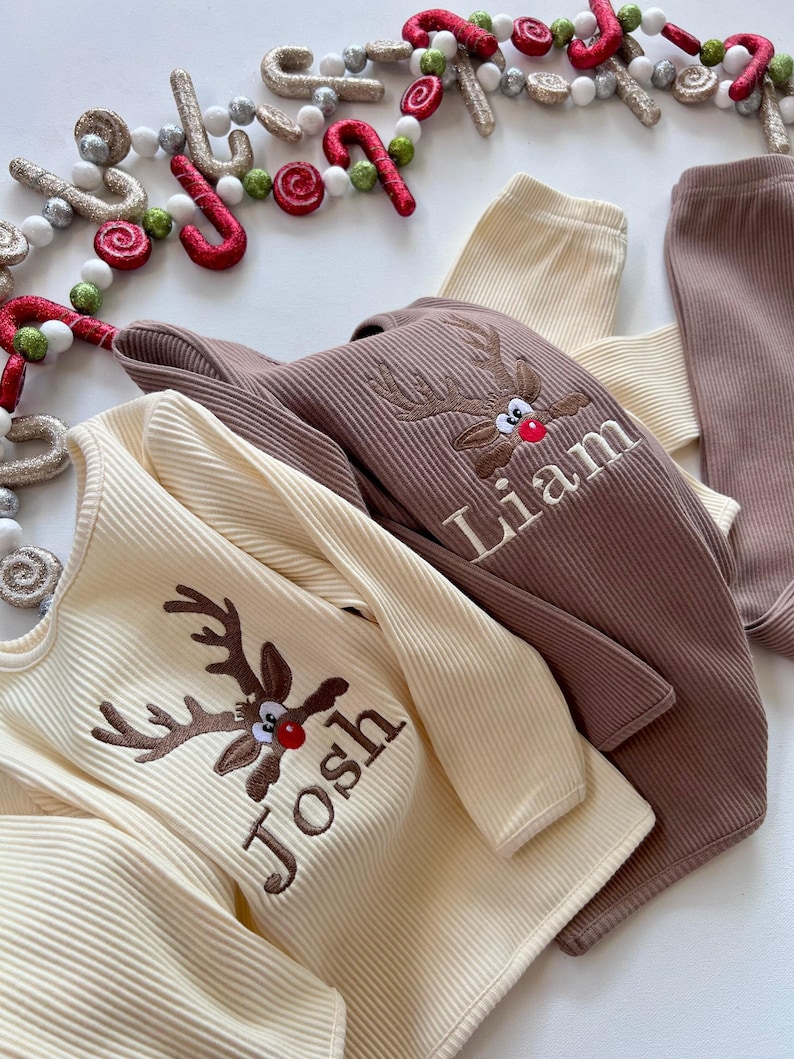May include: Cream and brown ribbed long-sleeved shirts and pants with embroidered reindeer faces and names. Festive holiday apparel with a Christmas theme. The text "Josh" and "Liam" are visible.