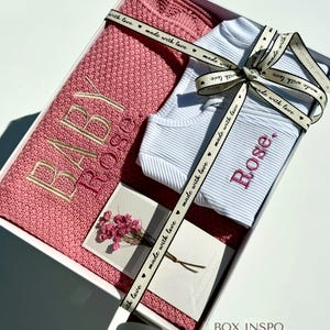 May include: A white gift box with a pink knitted blanket, a white romper with the name "Rose" embroidered on it, and a card with the text "made with love". The box is tied with a pink ribbon with the text "made with love" printed on it. The text "BOX INSPO" is printed on the bottom of the box, along with the text "French Rose blanket + White Romper, Font 4, Dahlia thread".