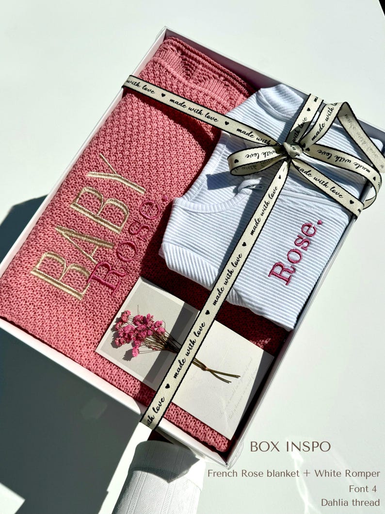 May include: A white gift box with a pink knitted blanket, a white romper with the name "Rose" embroidered on it, and a card with the text "made with love". The box is tied with a pink ribbon with the text "made with love" printed on it. The text "BOX INSPO" is printed on the bottom of the box, along with the text "French Rose blanket + White Romper, Font 4, Dahlia thread".