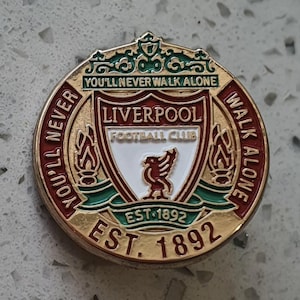 Puede incluir: Un pin circular dorado del Liverpool Football Club con borde rojo. El pin presenta el escudo del club, un Liver bird, y el texto "YOU'LL NEVER WALK ALONE" y "EST. 1892". Un artículo de colección.