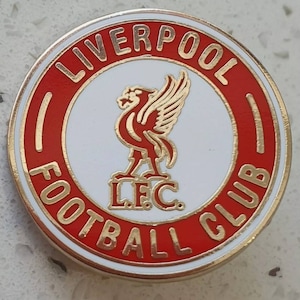 Puede incluir: Un pin circular rojo y dorado con las palabras "Liverpool Football Club" alrededor del borde. En el centro hay un círculo blanco con un pájaro rojo y las letras "L.F.C."