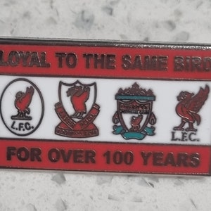 Puede incluir: Un pin de esmalte rojo, blanco y plateado con el texto "Loyal to the same bird for over 100 years". El pin presenta cuatro versiones diferentes del escudo del Liverpool Football Club.