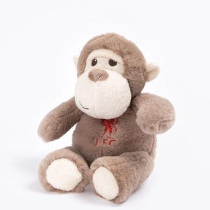 Może przedstawiać: Pluszowa zabawka małpka w odcieniach brązu i kremu, z uśmiechniętą twarzą i haftowanymi detalami. Małpka ma czerwone, haftowane logo na piersi. Zabawka jest przeznaczona do siedzenia.