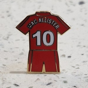 Puede incluir: Un pin de esmalte rojo y dorado con forma de camiseta de fútbol con el nombre "Mac Allister" y el número "10" en la espalda.