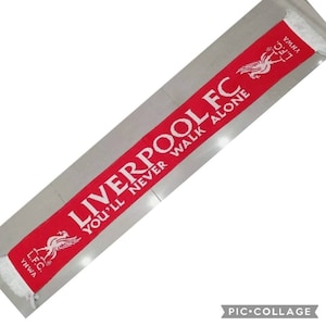 May include: Red and white scarf with the words "Liverpool FC You'll Never Walk Alone" and the Liverpool FC logo. The scarf has white fringe on both ends.