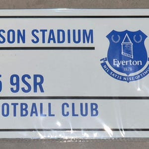 Puede incluir: Un letrero metálico blanco con texto negro y azul, con "HILL DICKINSON STADIUM", "BRAMLEY MOORE L5 9SR" y "EVERTON FOOTBALL CLUB". El logotipo del Everton está a la derecha. Una etiqueta de cartón azul está adjunta.