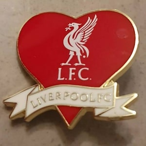 May include: A red heart-shaped pin with a white Liverpool Football Club logo. The logo features a bird with outstretched wings and the text "L.F.C." below. A white banner with the text "LIVERPOOLFC" is wrapped around the bottom of the heart.