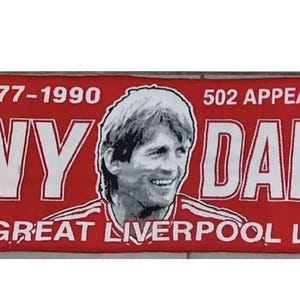 Puede incluir: Bandera roja con texto blanco y un retrato de Kenny Dalglish. La bandera dice "LIVERPOOL 1977-1990" y "502 APARICIONES, 169 GOLES". El texto "KENNY DALGLISH" se muestra de forma destacada, con "A TRUE GREAT LIVERPOOL LEGEND" debajo.