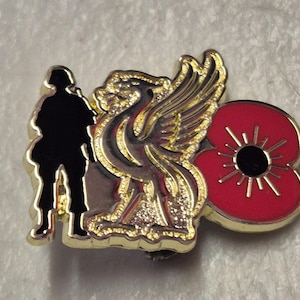 May include: A gold-coloured metal pin featuring a black silhouette of a soldier, a gold griffin, and a red poppy flower with a black centre. The pin has a shiny finish and is likely a commemorative item.