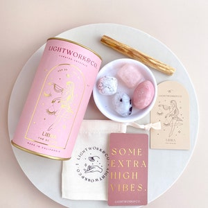 May include: Pink and gold Libra zodiac crystal kit with a pink cylinder container, a small white bowl with four crystals, a pink card with the text "Some Extra High Vibes. Lightwork&Co.", a pink card with a Libra zodiac symbol, and a wooden stick.