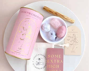 May include: Pink and gold Libra zodiac crystal kit with a pink cylinder container, a small white bowl with four crystals, a pink card with the text "Some Extra High Vibes. Lightwork&Co.", a pink card with a Libra zodiac symbol, and a wooden stick.