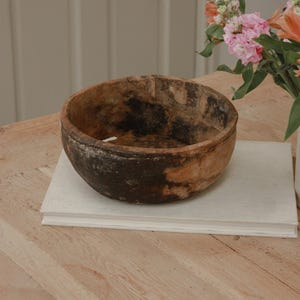 May include: A weathered wooden bowl, displaying dark brown and tan hues, rests on a stack of white books. The bowl's interior shows signs of use, enhancing its vintage charm. A bouquet of pink and orange flowers is in the background.
