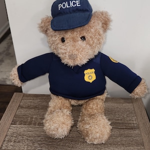 May include: A plush teddy bear dressed as a police officer. The bear wears a navy blue uniform with a matching cap that reads "POLICE". A yellow badge with "POLICE" is on the chest. The bear is light brown.