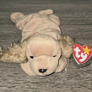 May include: A tan plush dog toy with floppy ears and a black nose. The toy has a red heart-shaped tag with the text "Ty" and a yellow star-shaped tag. The dog is lying down on a wooden surface.