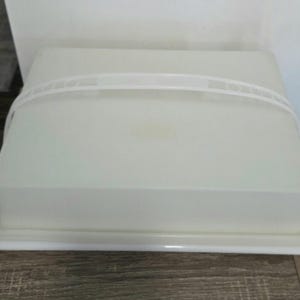 May include: A white rectangular cake carrier with a matching lid and a curved handle. The carrier is made of a translucent plastic material. The lid fits snugly over the base, providing a secure closure for transporting baked goods.