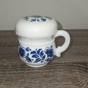 May include: A white ceramic shaving mug with a matching lid. The mug features a blue floral design and a curved handle. The lid also has a blue floral pattern. The mug is sitting on a wooden surface.