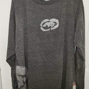 May include: Gray long-sleeve t-shirt with a white logo on the front. The logo is a stylized circle with a design inside. The shirt has a white graphic on the left sleeve.
