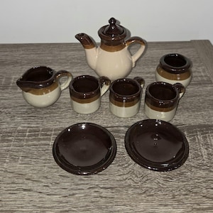 May include: A miniature ceramic tea set with a teapot, creamer, sugar bowl, cups, and saucers. The set features a two-tone design with a cream-colored base and brown accents. The teapot has a lid and a curved spout.